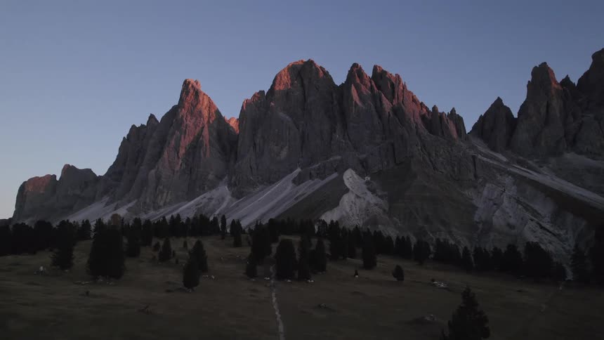 A mountain range with several sharp jagged peaks made of rock in varying shades of gray and brown is bathed in the warm golden light of the setting sun casting a gradient of blue and pink across the sky while a small cluster of trees and a building