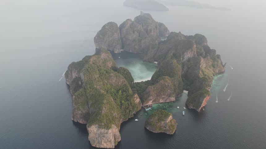 A birdseye view reveals a secluded island with dramatic cliffs and a hidden cove surrounded by the vast expanse of the ocean with a few boats dotting the water