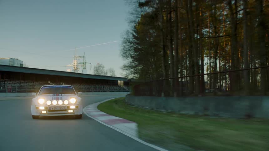 A vintage rally car with multiple frontmounted lights is driving on a racetrack