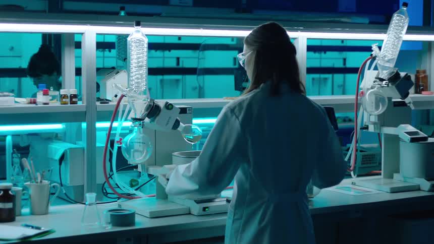 Woman dressed in a white lab coat and safety glasses works in a bluetoned laboratory filled with equipment