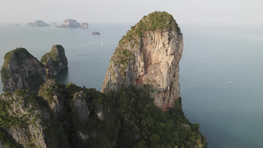 A towering limestone cliff covered in lush vegetation dominates the landscape with smaller islands and boats visible in the distance