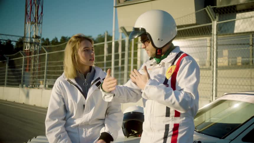 A man in a helmet is talking to a blonde woman making gestures with his hands and both are wearing white clothes next to a car