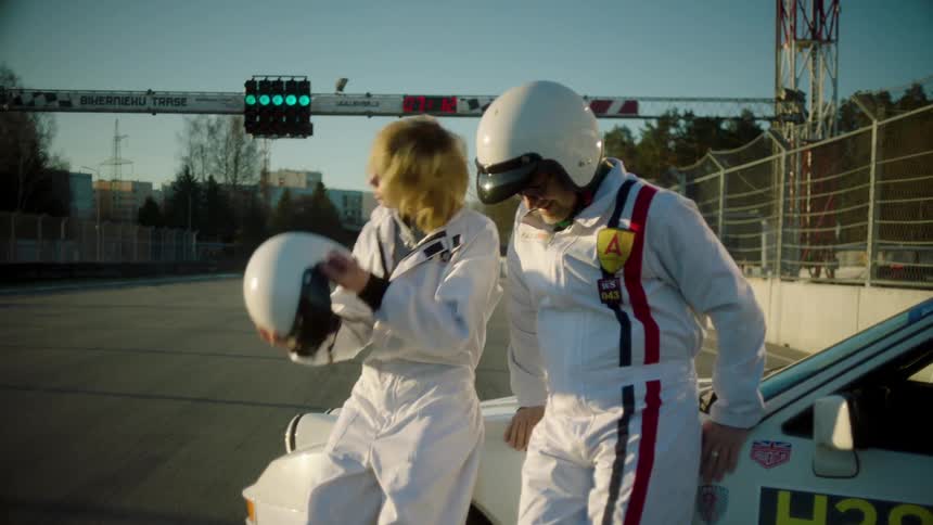 A clip of a male and a female discussing beside a car on a race track