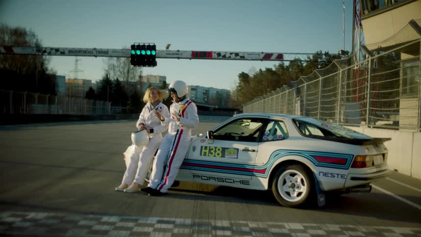 Woman taking off her helmet next to a man and both in sportswear next to a car on a race track