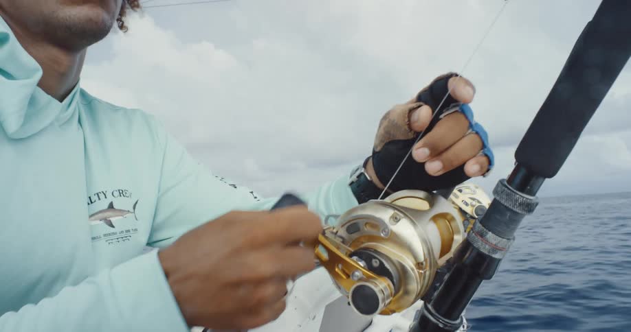 A persons hands turning the reel handle of a fishing rod that is fixed to a boat moving in a blue sea