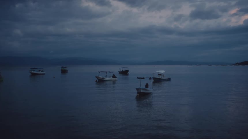 A cluster of small boats bob gently on the surface of a still lake at dusk under a sky shrouded in dark ominous clouds