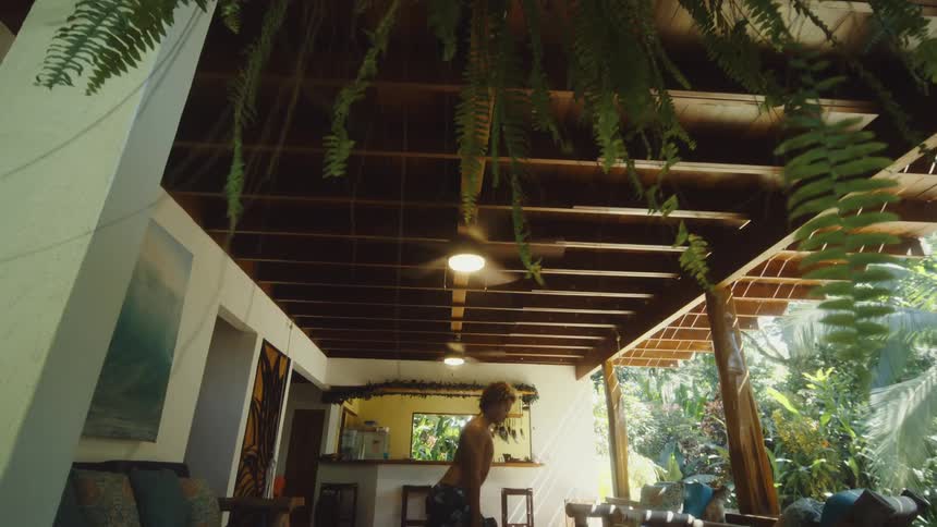 A person performs a lunge exercise in an openair living area with wooden furniture tropical plants and a kitchen in the background