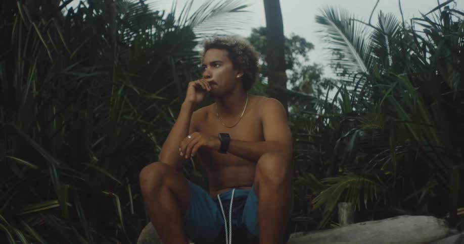 A shirtless person with curly hair sits on a log in a dense jungle surrounded by tall palm fronds and tropical vegetation