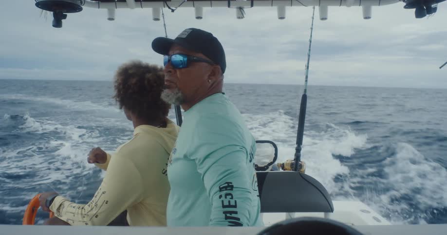 Two men are on a boat looking around as it moves through the water