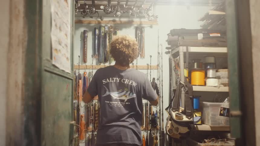 A young man in a salty crew shirt enters a small storage room filled with fishing gear selects a lure from the wall and then leaves the room
