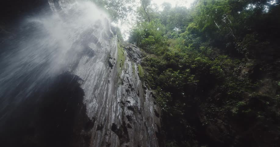 A tall misty waterfall cascades down a steep rocky cliff surrounded by lush green foliage