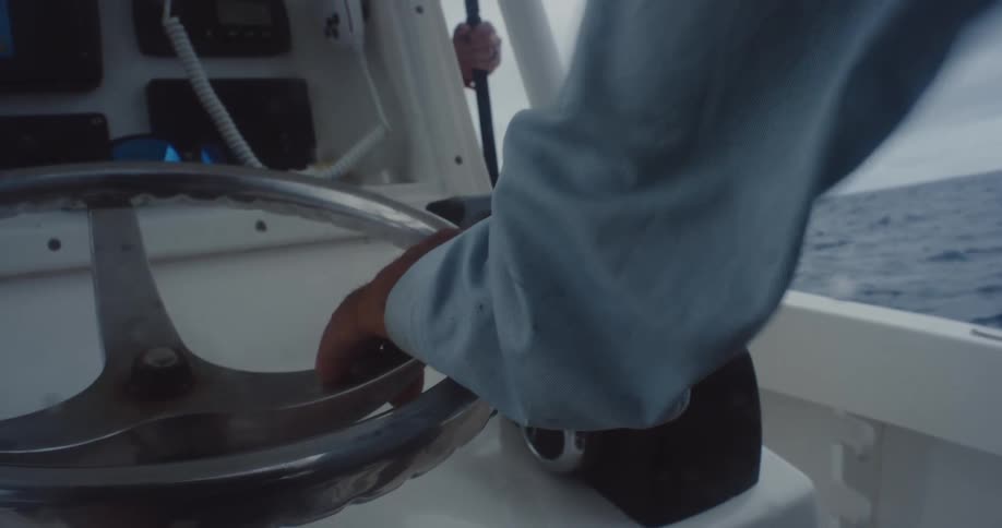 The hand of a person wearing a long blue sleeved outfit moving in the wind and is moving the steering wheel of a boat that is sailing on the sea