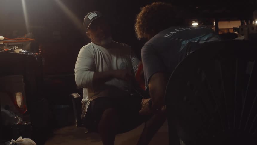 Two men sitting with one of them reeling in a fishing line