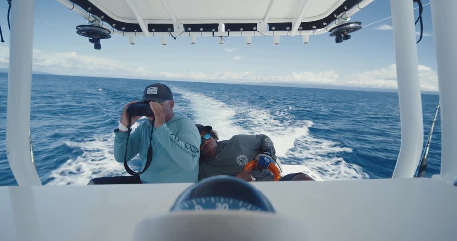Son naps while his father uses binoculars and steers their boat across the ocean surface on a cloudy day