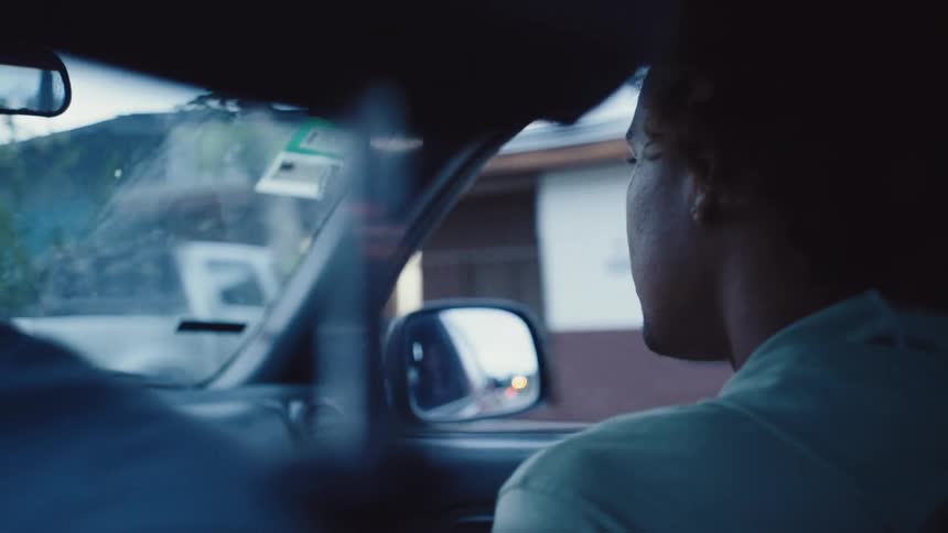 Two men in a car are going down a street past houses and cars
