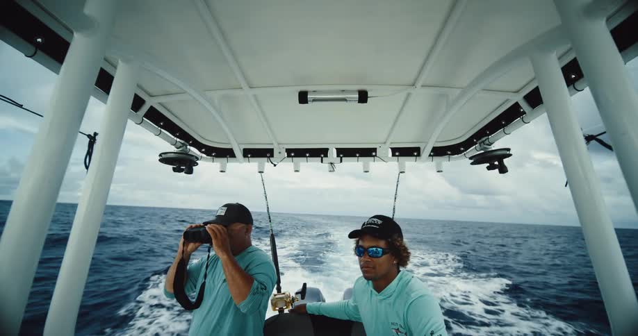 Two men in dark caps and blue shirts are on a white boat sailing in the ocean and one of them is using binoculars to look at the place
