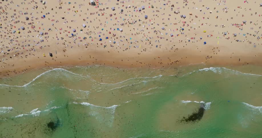 An aerial view of a crowded beach shows numerous people umbrellas and beach accessories scattered across the sand with waves gently crashing onto the shore