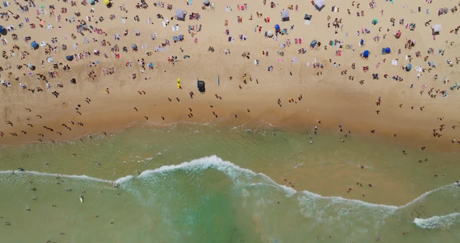 An aerial view shows the beach covered with people lying on towels and blankets some under umbrellas enjoying the sun on a summer day