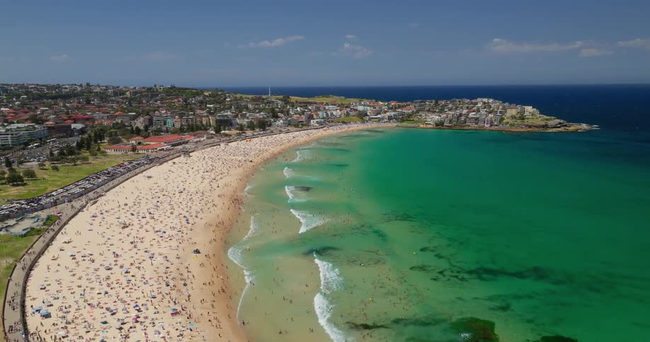 A panoramic view captures bondi beach on a sunny day showing thousands of sunbathers dotting the golden sand while the turquoise water laps at the shore and highrise buildings line the coast
