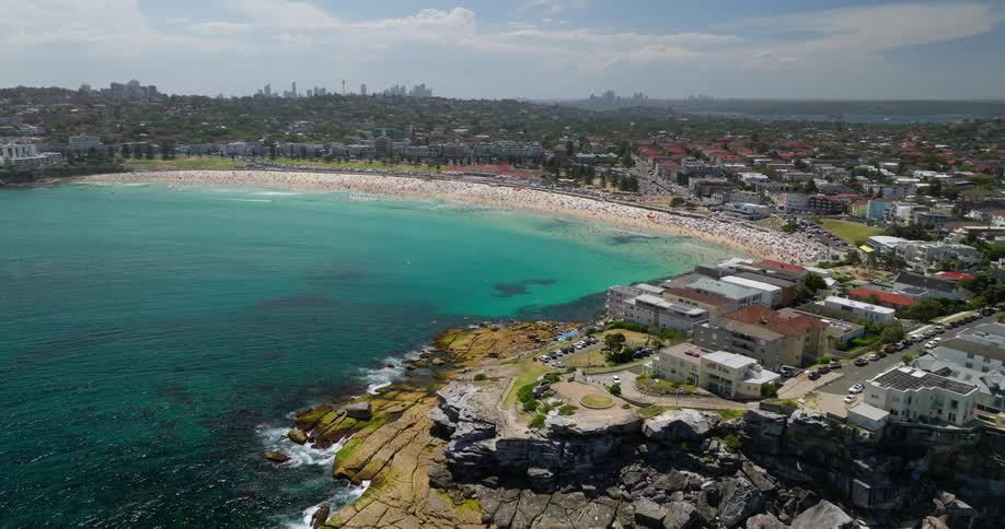 An aerial view of a coastal city featuring a sandy beach with turquoise waters rocky cliffs with waves crashing against them and a densely built urban area in the background