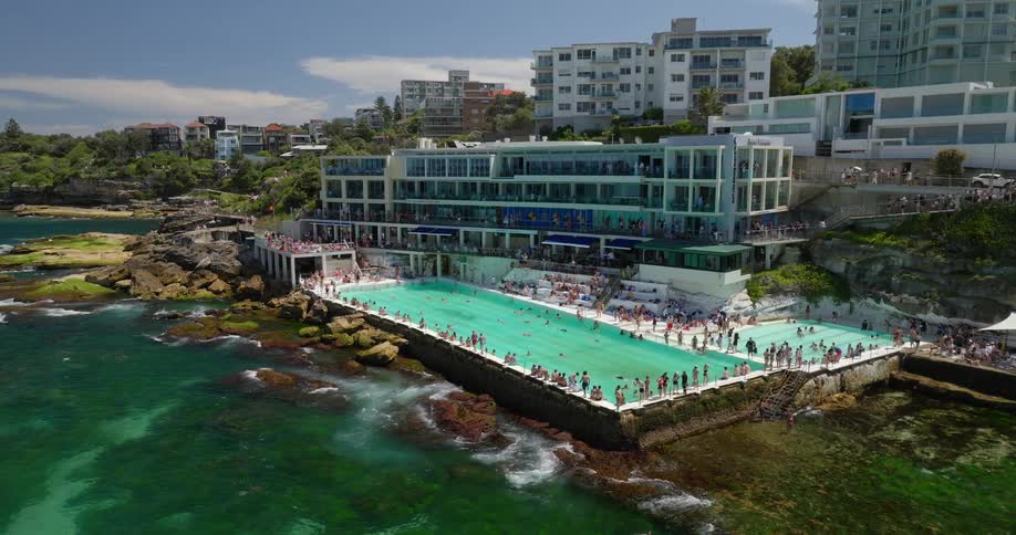 People enjoy coastal swimming pools in a town bordered by woods on a cloudy day