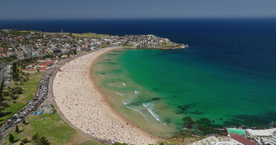 An aerial view shows bondi beach with many people lying on the golden sand and the water is a bright turquoise color with waves sparkling in the sun
