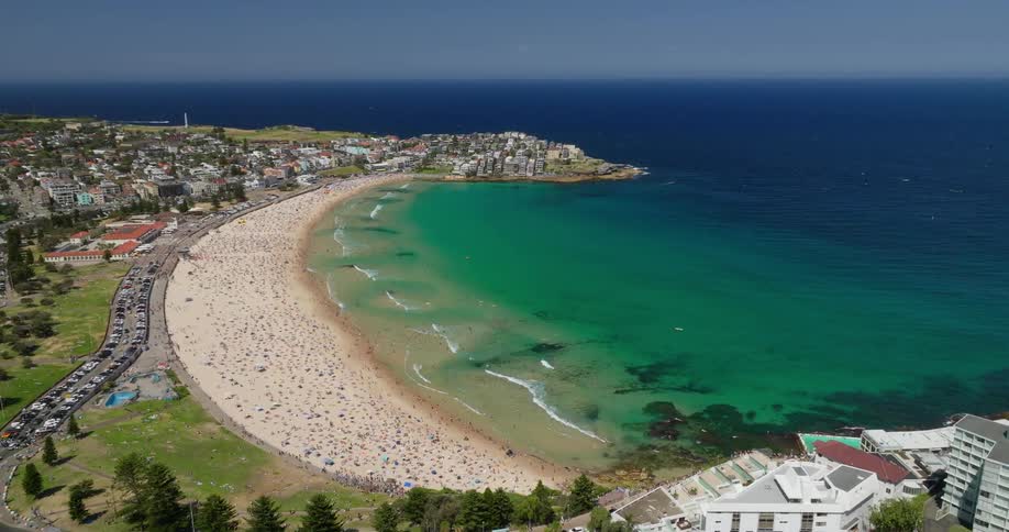 Aerial view of bondi beach on a sunny day showing thousands of sunbathers dotting the golden sand while the turquoise water laps at the shore and highrise buildings line the coast