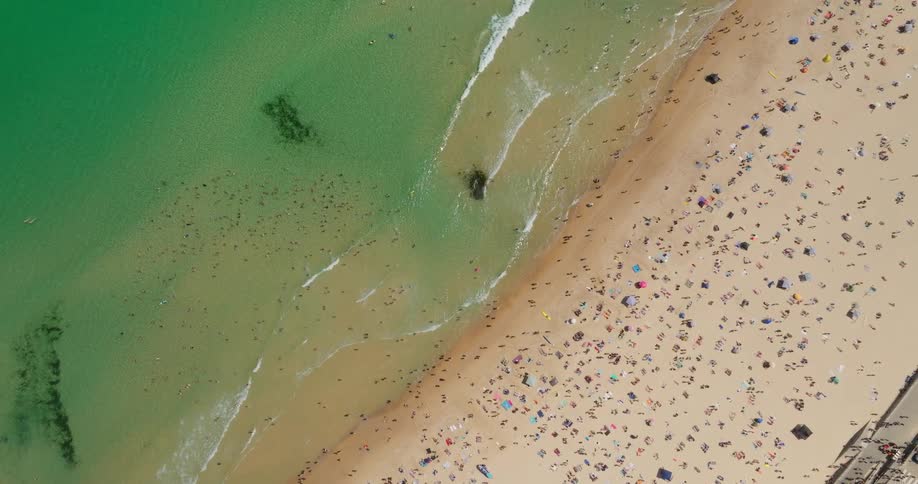 An aerial view of a beach full of people on the sand and some waves in the water breaking on the shore