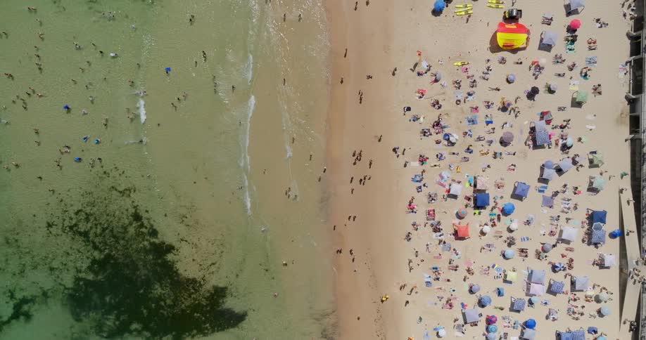 A crowded beach stretches along the coastline with sunbathers scattered across the sand and swimmers enjoying the clear water