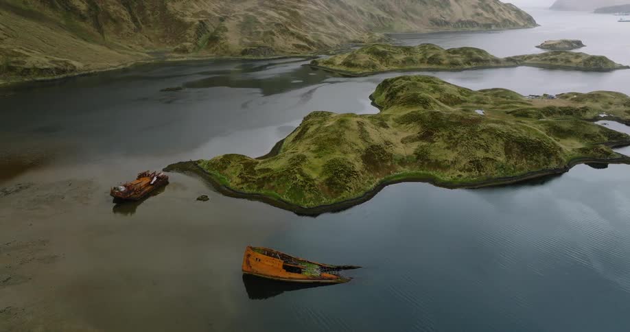 A rugged green island is surrounded by calm waters with two rusted shipwrecks partially submerged near its shores