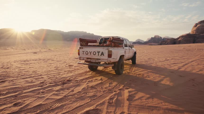 A white pickup truck driving in a desert with mountains in the background and sunlight coming from the left