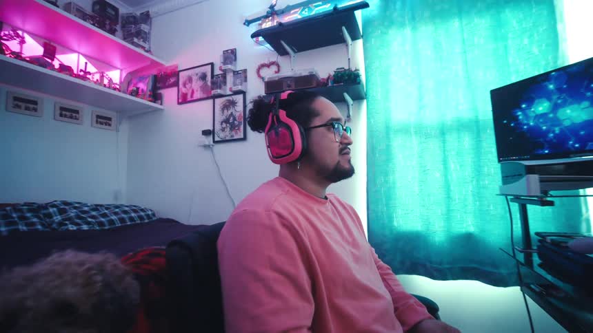 A man with glasses and pink headphones is sitting at his computer playing a video game while his dog lays on the bed behind him