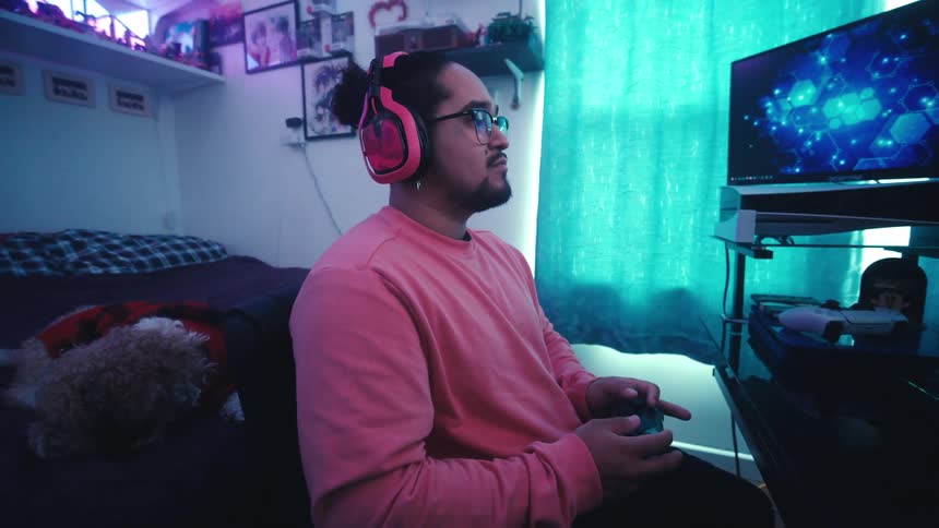 A man wearing a pink sweater and headphones playing videogames in his room