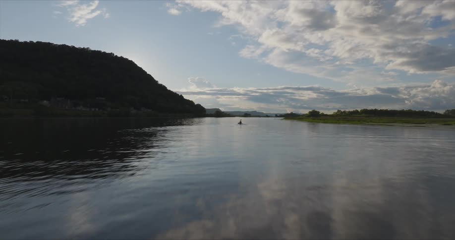 Person paddles a kayak through the water between a field and a mountain on a cloudy day
