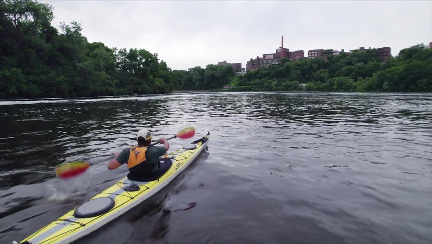 A kayaker paddles a yellow kayak down a river with a forested bank on one side and a city skyline visible in the background
