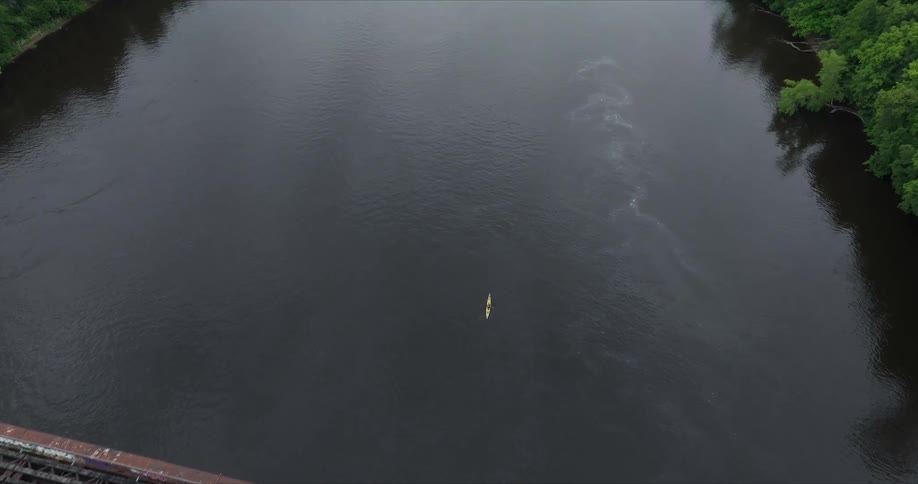 An aerial shot pans from a rusty railroad bridge over a river towards a single kayaker paddling downstream