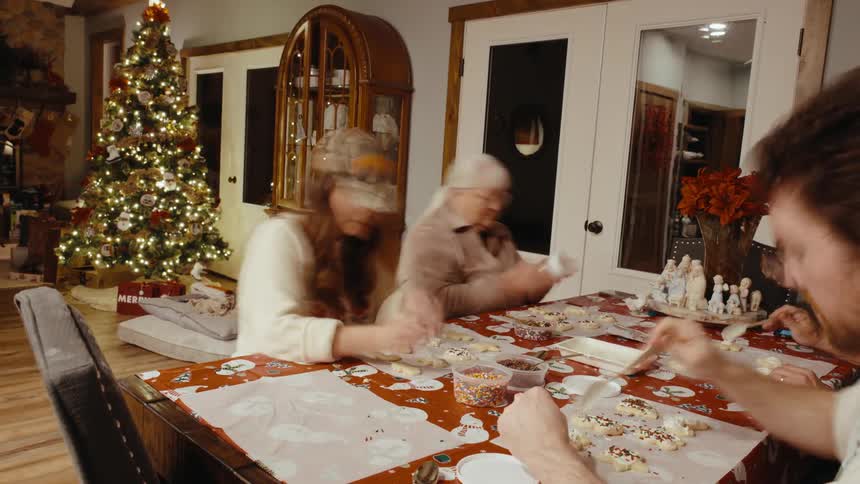 Family gathered around a table decorated with a christmas themed tablecloth preparing decorative cookies