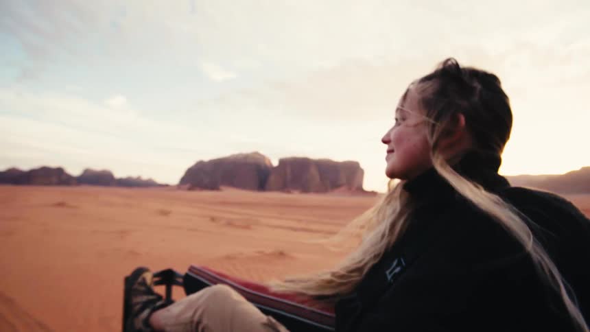 Woman sits on top of a moving vehicle enjoying the vastness of a reddish desert at dusk