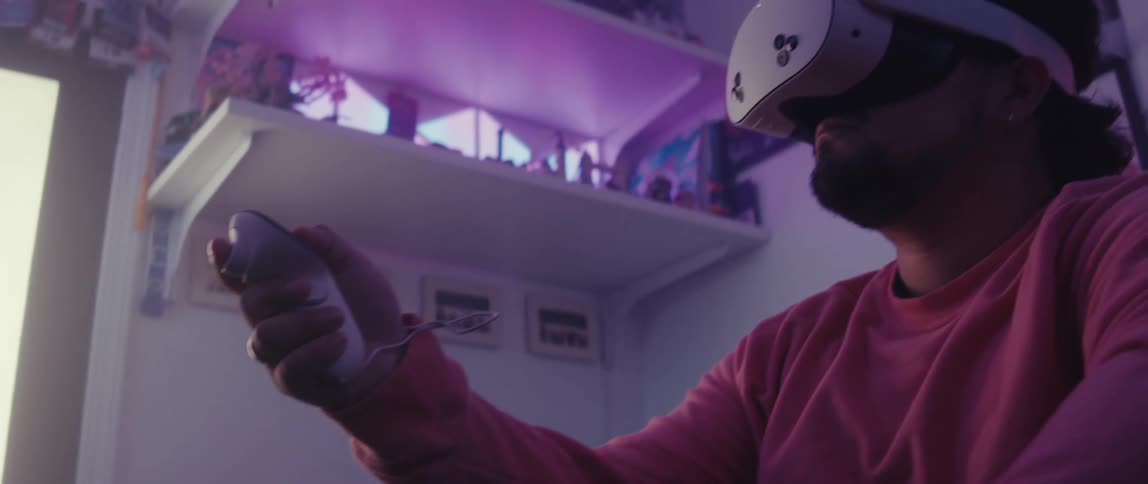 A man in a pink shirt wearing a virtual reality headset and holding a controller immersed in a vr experience with dim lighting and a purple glow in the background