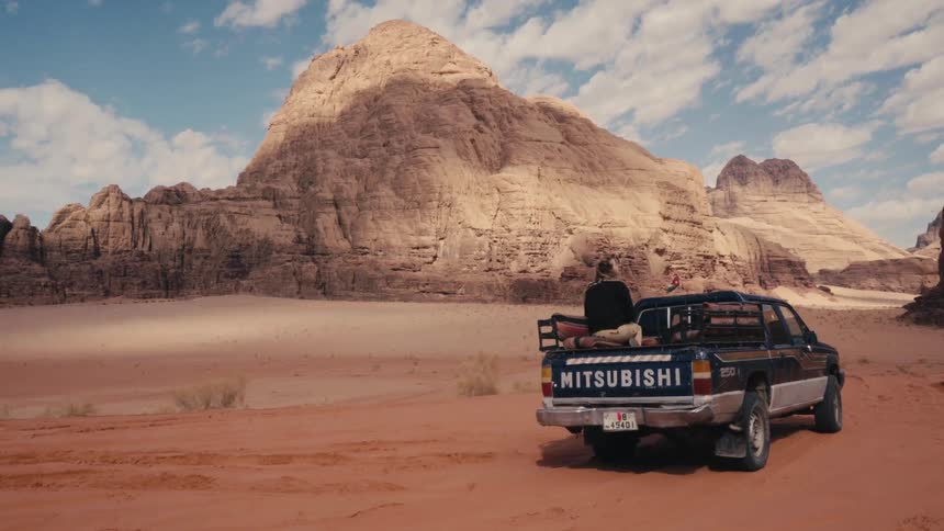 Woman sitting in the back of a pickup truck parked in arid terrain near rocky hills on a windy day looks around