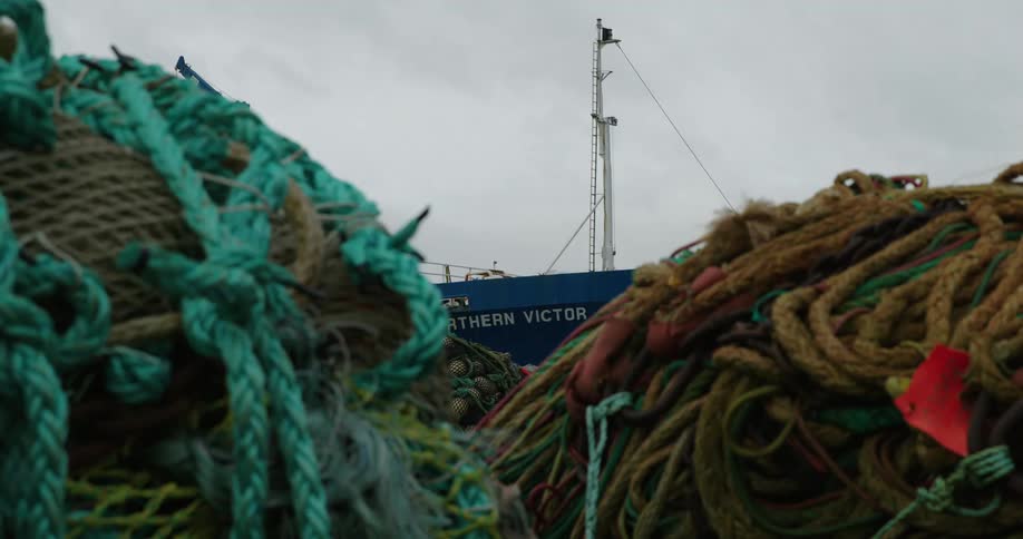 A large pile of colorful fishing nets and ropes sits in front of a blue ship named northern victor