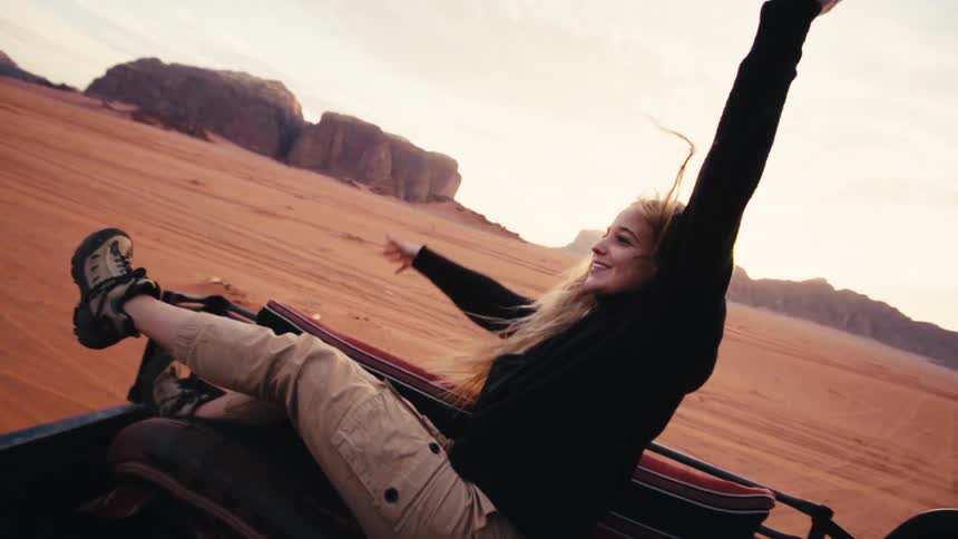 A woman playfully leans back as she rides in the back of a truck through a desert area