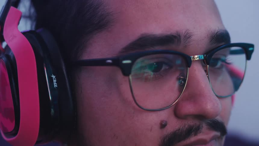 A close up shot shows a man wearing glasses and pink headphones looking intently ahead