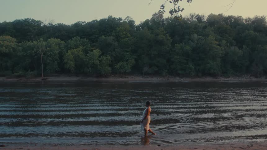 A woman wearing a white dress walks with her feet submerged in the shallow river water