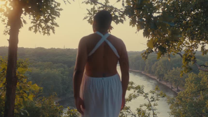 Woman walks to a point overlooking a river passing through the woods and looks around on a sunny day