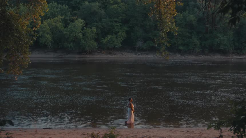 A woman in a light colored dress stands in a river at the edge of a sandy bank surrounded by trees and forest