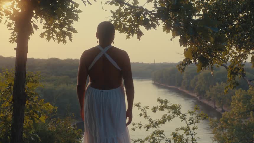 A person in a white dress stands on a hilltop overlooking a serene river at sunset framed by lush green trees