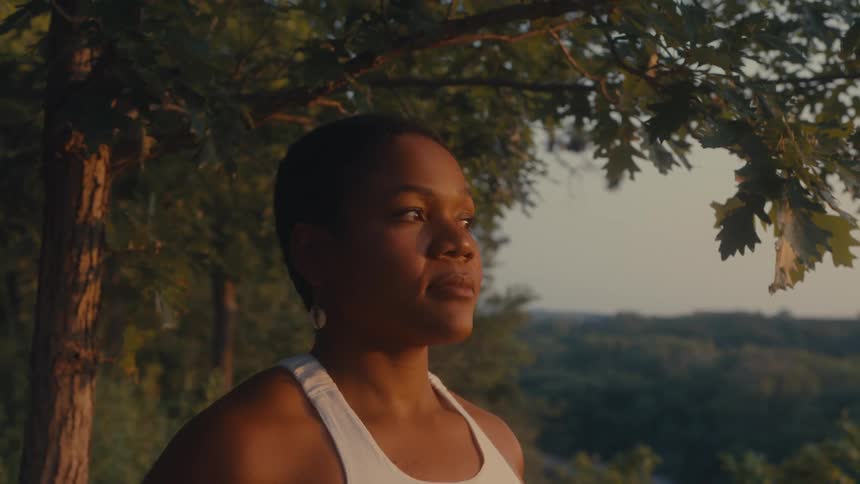 A woman wearing a white tank top standing next to a tree in the wilderness at sunset
