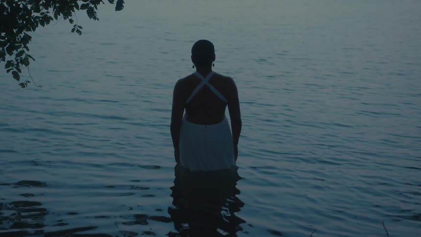 A woman in a white dress stands waistdeep in calm water facing away from the camera