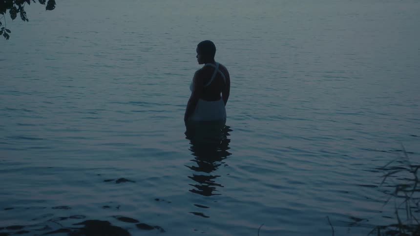 A person in a white dress stands waistdeep in calm water facing away from the camera